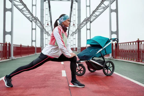 Tiro de comprimento total de jovem motivada esticando as pernas, de pé na ponte com uma carruagem de bebê, preparando-se para uma corrida em um dia nublado na cidade — Fotografia de Stock