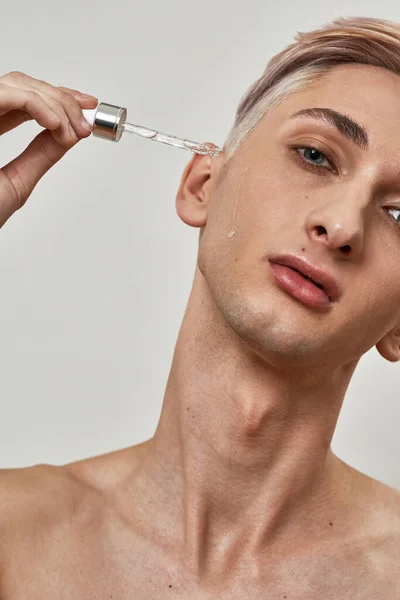 Portrait of shirtless young transgender looking at camera holding pipette and applying hyaluronic acid from the bottle, posing isolated over light background — Stok Foto