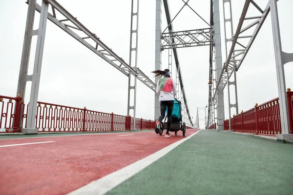 Αθλητική νεαρή μητέρα τρέχει με ένα καροτσάκι μωρού σε μια γέφυρα στην πόλη σε μια συννεφιασμένη μέρα — Φωτογραφία Αρχείου