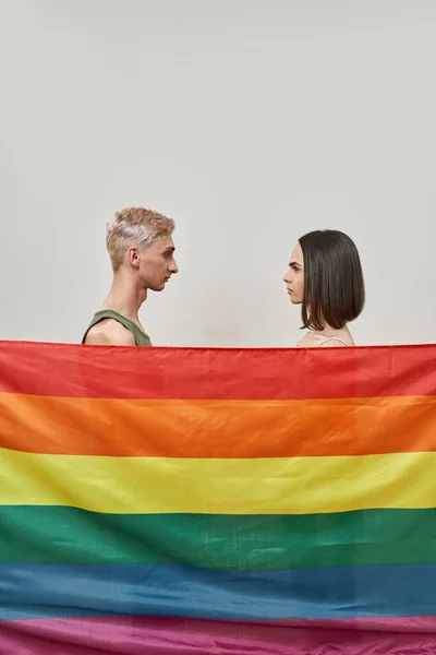 Вид сбоку молодой трансгендерной пары, стоящей за разноцветным радужным флагом ЛГБТ и смотрящей друг на друга, позируя вместе на сером фоне — стоковое фото