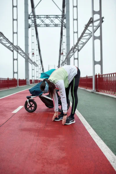 在这个多云的城市里，一个年轻的女运动员站在桥上，提着婴儿车，全身伸展着身子 — 图库照片