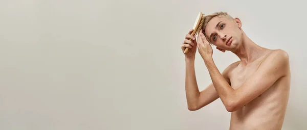 Jovem transgênero escovar seu cabelo curto luz usando pente de cabelo, olhando para longe enquanto posando isolado sobre fundo claro — Fotografia de Stock