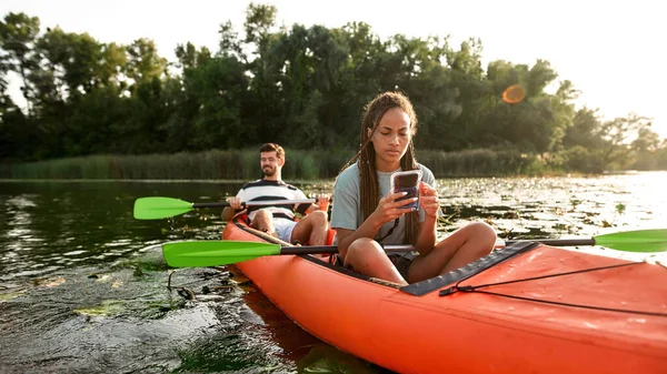 Porträt einer jungen Frau, die ihr Smartphone in der Hand hält und SMS schreibt, während sie Zeit mit ihrem Freund im Freien mit dem Kajak auf dem Fluss verbringt — Stockfoto
