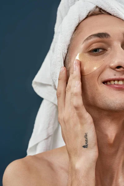 Polovina obličeje detailní záběr šťastný mladý transsexuál s ručníkem zábal nanášení pod oko gel náplasti dávat pozor, usmívat se na kameru izolované přes tmavé pozadí — Stock fotografie