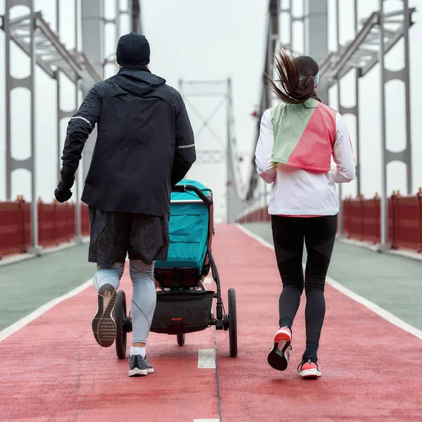 Visão traseira do casal esportivo, pais jovens correndo juntos com uma carruagem de bebê em uma ponte na cidade em um dia nublado — Fotografia de Stock