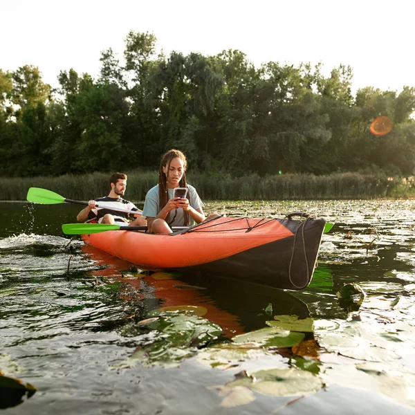 Junge Frau hält Smartphone und SMS in der Hand, während sie Zeit mit ihrem Freund im Freien mit dem Kajak auf dem Fluss verbringt — Stockfoto