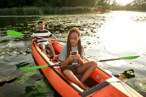 Mladá žena pomocí svého chytrého telefonu, zatímco tráví čas se svým přítelem venku, kajak na řece — Stock fotografie