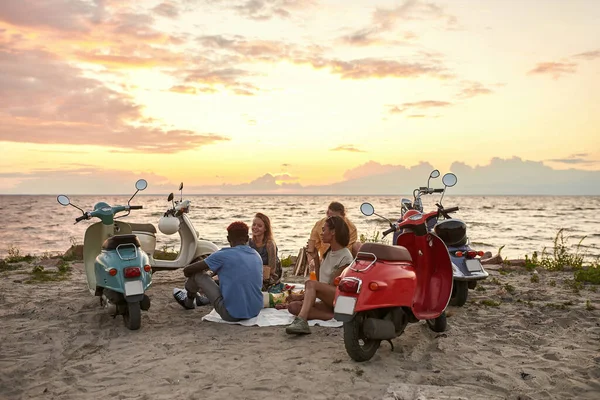 Jóvenes caucásicos, motociclistas disfrutando de la vista mientras hacen picnic en la playa al atardecer — Foto de Stock