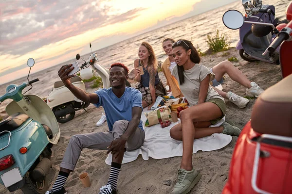 Jóvenes alegres y diversos sonriendo mientras se toman selfie, haciendo picnic, pasando tiempo juntos en la playa — Foto de Stock