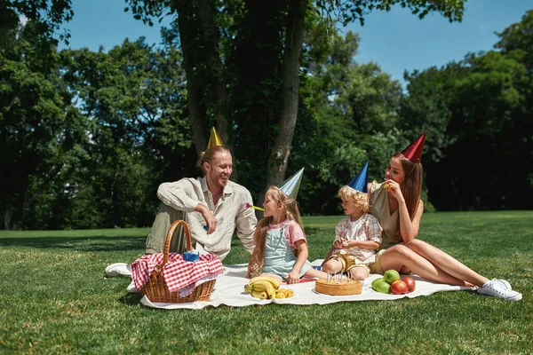 Glada familjen bär fest hattar, firar barn födelsedag, har picknick i parken på en sommardag. Barn som blåser födelsedagsvisslingar — Stockfoto