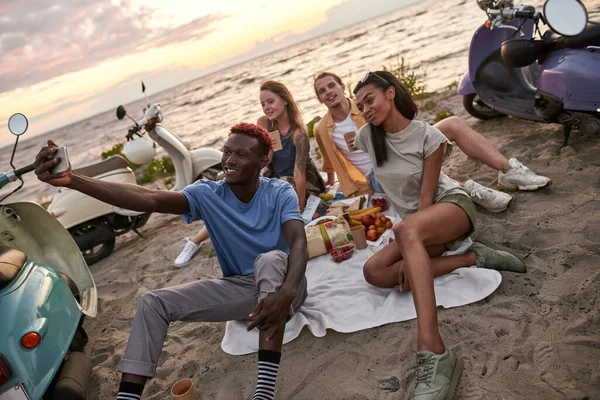 Felices jóvenes diversos sonriendo mientras se toman selfie, haciendo picnic, pasando tiempo juntos en la playa — Foto de Stock