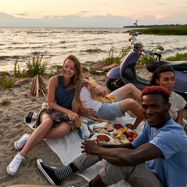 Jóvenes alegres sonriendo a la cámara mientras disfrutan de un picnic, sentados en la playa al atardecer — Foto de Stock