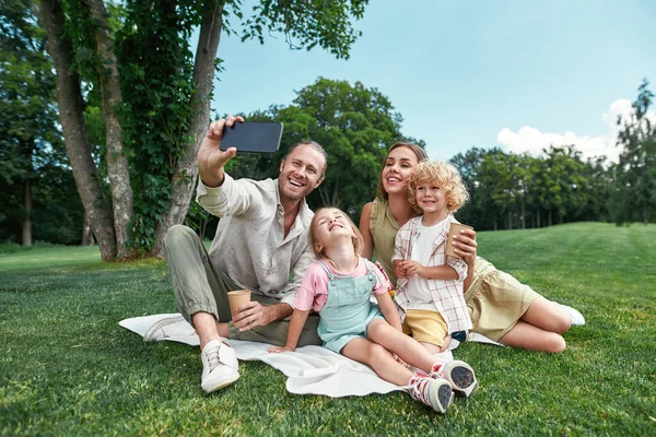 Piękna rodzina spędzać czas razem na świeżym powietrzu, za pomocą smartfona podczas robienia selfie, o piknik w przyrodzie w letni dzień — Zdjęcie stockowe