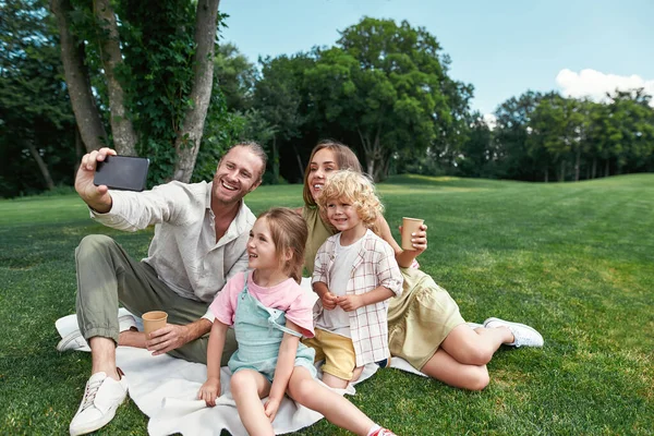 Alegre familia pasar tiempo juntos al aire libre, utilizando el teléfono inteligente mientras se toma una selfie, teniendo picnic en la naturaleza en un día de verano — Foto de Stock