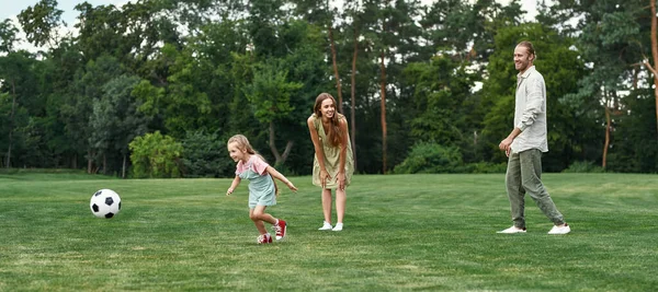 Holčička honí míč. Mladá rodina hraje fotbal na trávníku v parku v letní den — Stock fotografie