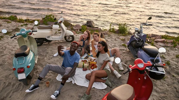 Jóvenes, ciclistas de scooter tomando selfie mientras hacen picnic en la playa, pasando tiempo juntos fuera de la ciudad — Foto de Stock