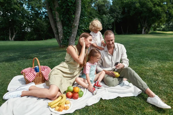 Härlig familj som tillbringar tid utomhus. Föräldrar med små barn tittar på bilder de tog samtidigt ha picknick i naturen på en sommardag — Stockfoto