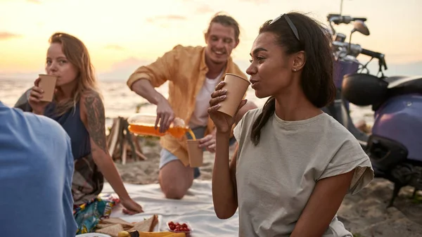 Alegres jóvenes amigos de vacaciones disfrutando de la vista mientras hacen un picnic en la playa al atardecer — Foto de Stock