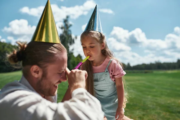 Cute girl wearing party hat, blowing whistle in father ear while celebrating his birthday in the park on a summer day — Stock Photo, Image