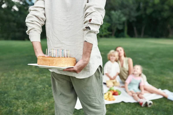 Close up of father holding and hiding a birthday cake for his child. Family relaxing, having picnic in the park outdoors on a summer day