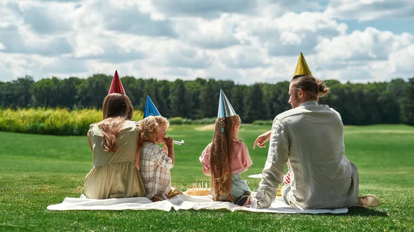 Vista trasera de la encantadora familia con sombreros de fiesta, celebrando el cumpleaños de los niños, haciendo un picnic en el parque en un día de verano — Foto de Stock