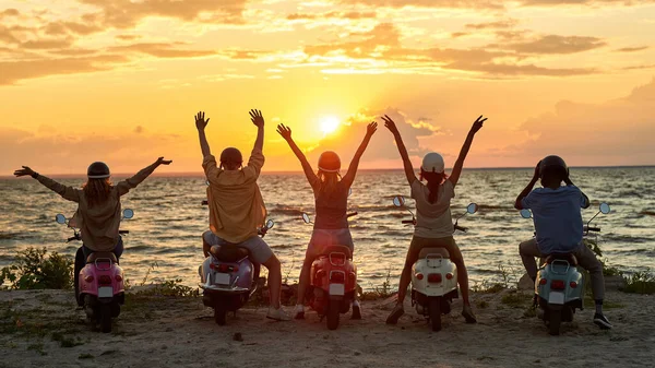 Vista posterior de los jóvenes amigos, motociclistas levantaron las manos mientras estaban de pie con scooters en la playa y disfrutando de la puesta de sol juntos — Foto de Stock