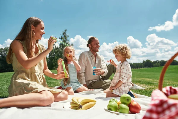 Två små barn blåser såpbubblor medan spendera tid med sina föräldrar, familj har picknick i naturen på en sommardag — Stockfoto