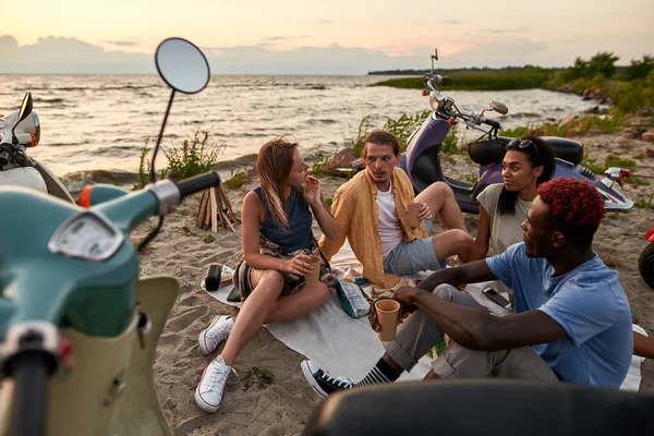 Jóvenes relajados disfrutando de un picnic, hablando mientras están sentados en la playa al atardecer — Foto de Stock