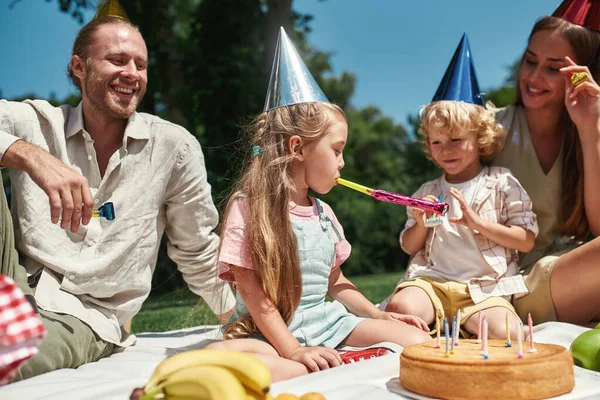 Barn i partyhattar har roligt, blåser födelsedagsvisselpipa samtidigt som de har fest med föräldrar i parken på en solig dag — Stockfoto