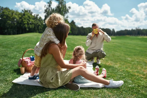 Bakåt syn på ung mamma med små barn poserar medan pappa tar en bild av dem med hjälp av filmkamera i naturen på en sommardag — Stockfoto