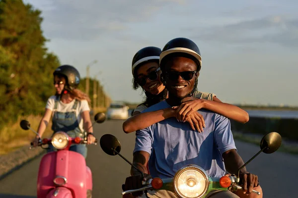 Smiling young diverse couple wearing protective helmets and sunglasses, riding vintage moto scooter together with friends outside the city at sunset — Stock Photo, Image