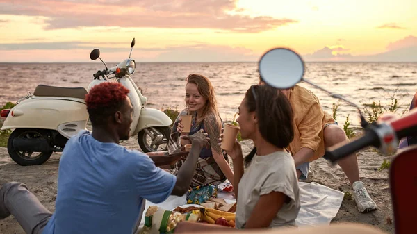 Jóvenes alegres, motociclistas disfrutando de la vista mientras hacen picnic en la playa al atardecer — Foto de Stock