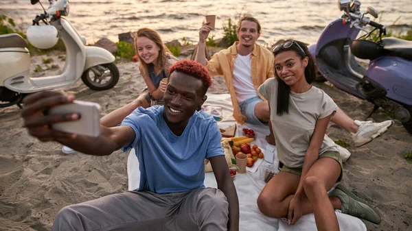 Kaum muda yang beragam bersenang-senang, berfoto selfie selama piknik yang menyenangkan di pantai dengan buah-buahan dan minuman yang sehat — Stok Foto