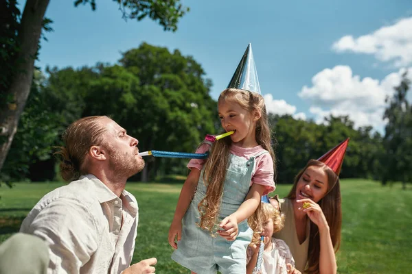 Gelukkige vader draagt feestmuts, blaast samen met kinderen op fluit, viert verjaardag in het park op een zomerdag — Stockfoto