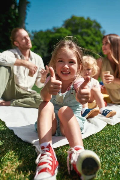 Gelukkig klein meisje tonen duimen tot camera terwijl ontspannen met familie buiten, picknick in de natuur op een zomerse dag — Stockfoto