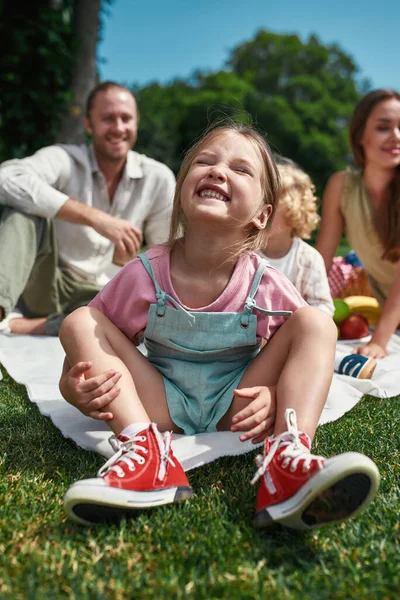 Linda niña sonriendo a la cámara mientras se relaja con la familia al aire libre, teniendo picnic en la naturaleza en un día de verano — Foto de Stock