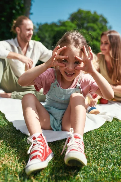 Zabawna dziewczynka robi miny przed kamerą, relaksując się z rodziną na świeżym powietrzu, organizując piknik w przyrodzie w letni dzień — Zdjęcie stockowe