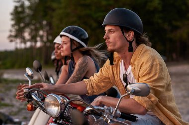 Kasklı neşeli genç motosikletli adam şehir dışında arkadaşlarıyla vakit geçiriyor, mobiletin üzerinde oturuyor ve gün batımını seyrediyor.