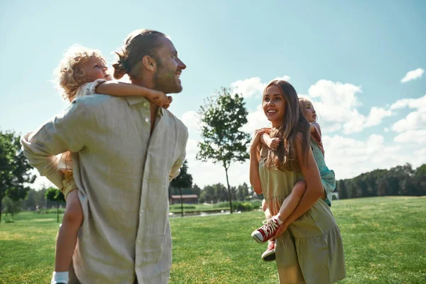 Yeşil yaz parkında birlikte yürürken küçük çocuklarını sırtında taşıyan genç aileler. — Stok fotoğraf