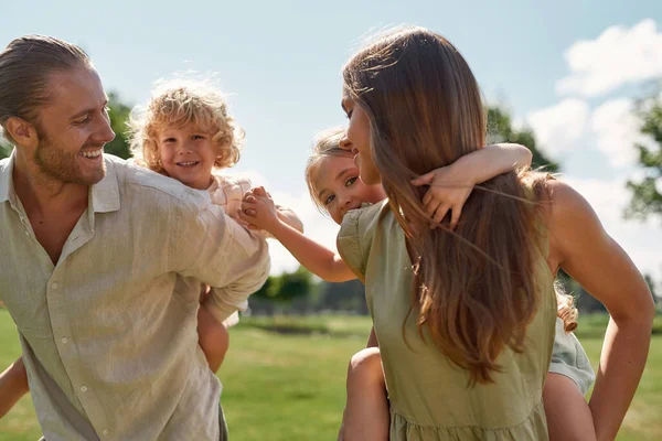 Små barn, pojke och flicka njuter av piggyback rida på baksidan av sina föräldrar. Familjen tillbringar tid tillsammans i grön sommarpark — Stockfoto