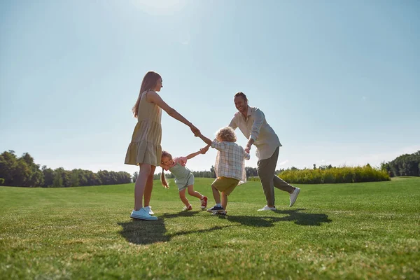 Active young parents spending time together, holding hands in circle with their two little kids, boy and girl in green park on a summer day