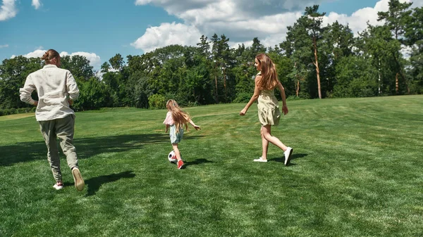 Familia joven activa jugando al fútbol junto con su hija pequeña en el campo de hierba en el parque en un día de verano — Foto de Stock
