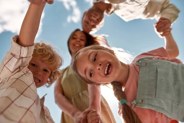 Retrato de dos pequeños niños alegres tomados de la mano con sus padres y mirando hacia abajo a la cámara con el cielo azul por encima de ellos —  Fotos de Stock