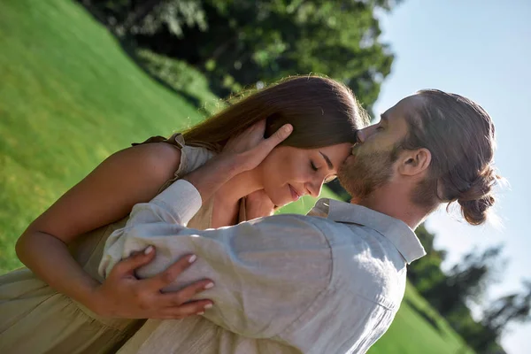 Soțul afectuos sărutându-și frumoasa soție în frunte în timp ce pozează împreună în aer liber, stând în parc — Fotografie, imagine de stoc