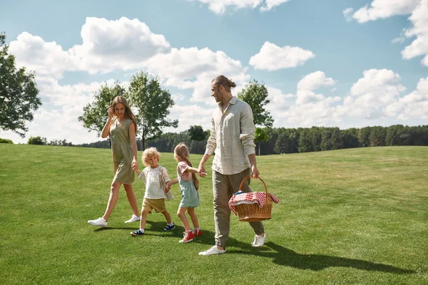 Plan complet de jeunes parents heureux avec deux petits enfants marchant dans un parc verdoyant, prêts à pique-niquer dans la nature un jour d'été — Photo