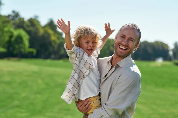 可爱的爸爸对着镜头笑着，夏天和他在草地上玩耍的时候抱着他的孩子。小男孩抬起胳膊，看上去很高兴 — 图库照片