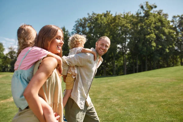 Unge far ler mot kameran. Små barn njuter av en åktur på baksidan av sin mamma och pappa. Familjevandring i grön sommarpark — Stockfoto