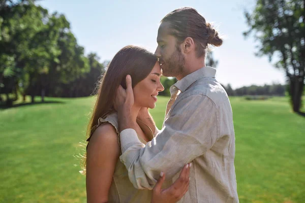 Affectueux homme embrasser jeune femme dans le front alors qu'ils posent ensemble à l'extérieur, debout dans le parc — Photo