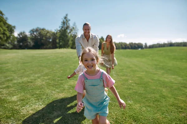 Vidám aranyos kislány néz izgatott a kamera, szórakozás együtt szüleivel és bátyjával, fut a zöld parkban egy nyári napon — Stock Fotó