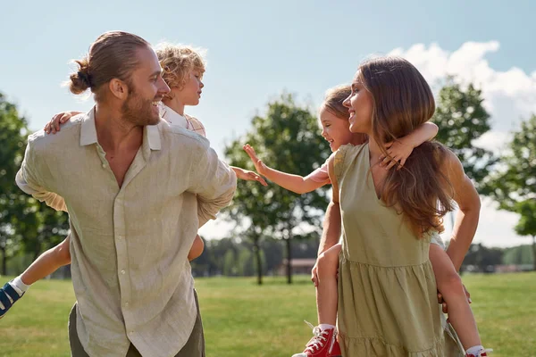 Genç ebeveynler küçük çocuklarına sırtına bindirirken birbirlerine gülümserler. Aile yeşil yaz parkında yürüyor. — Stok fotoğraf